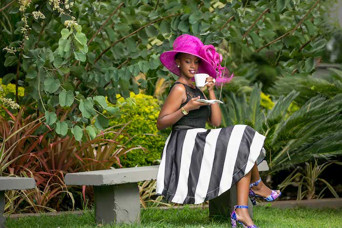 hot pink fancy hat, worn by woman, sitting on a stone bench in a garden, wearing black and white striped dress, and multicolored high heel sandals, vintage garden party dress code