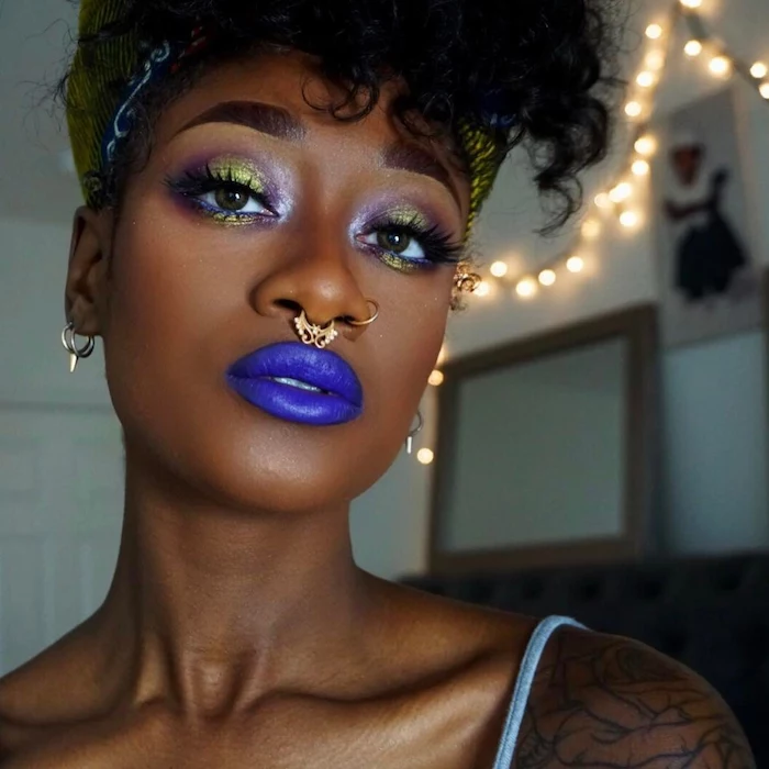 dark blue lipstick, worn by black woman, with eye shadow in yellow, purple and white, black eyeliner and fake lashes, festival makeup, nose rings and earrings