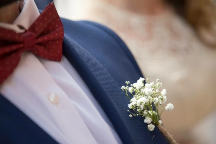 close up of a man's boutonniere, small bunch of baby's breath white flowers, mens summer wedding attire, dark blue suit jacket, worn with a white shirt and red bowtie