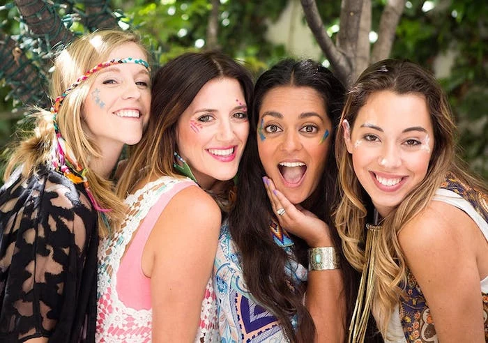 group of friends, made up of four women, smiling and posing for a photo, wearing boho style clothing, and face paint in different patterns and colors