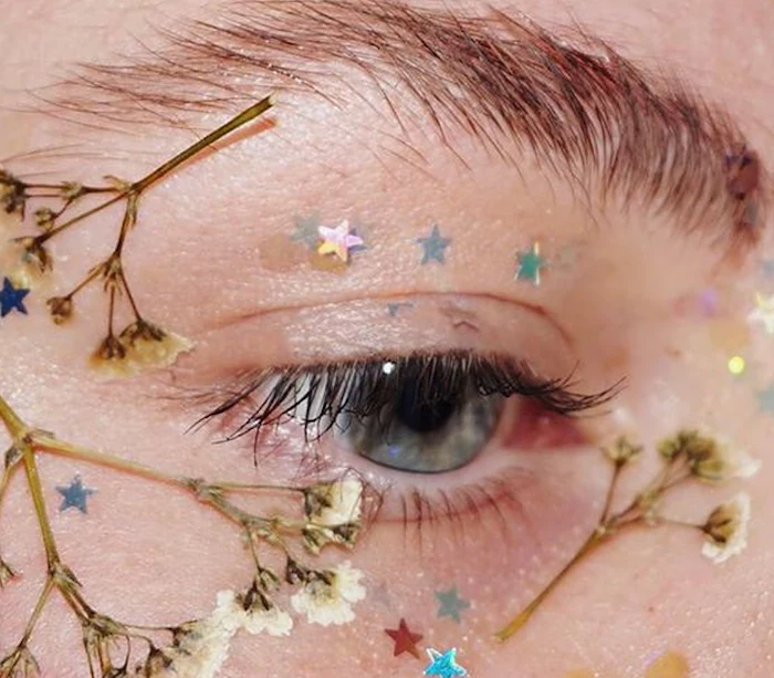 pressed tiny white dried flowers, stuck around a blue eye, seen in close up, with iridescent glitter stars, festival makeup, bushy brunette eyebrow