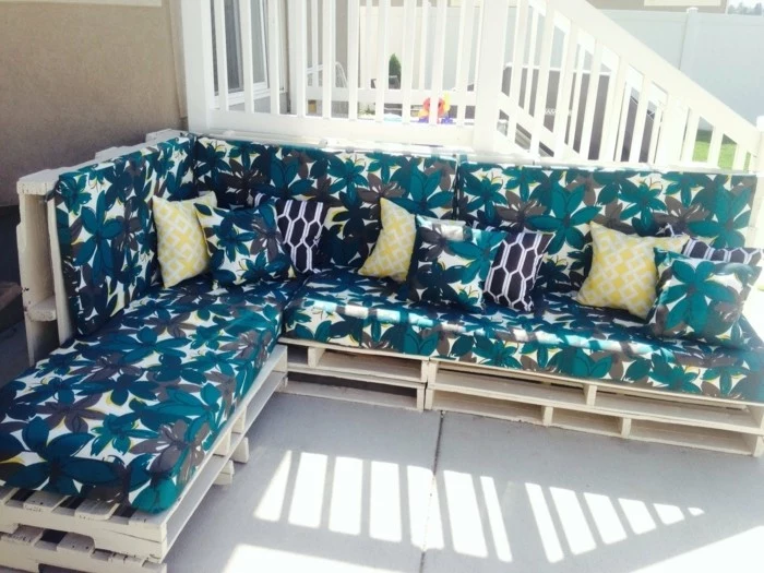 porch near a white staircase, with a big corner couch, made from pale wooden pallets, foam mattresses in blue, grey and white floral pattern
