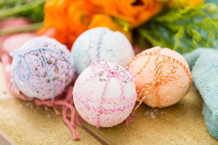 set of four bath balls, in pale purple, light pink and blue, and pale orange, placed on a wooden surface, and decorated with striped string, in corresponding colors