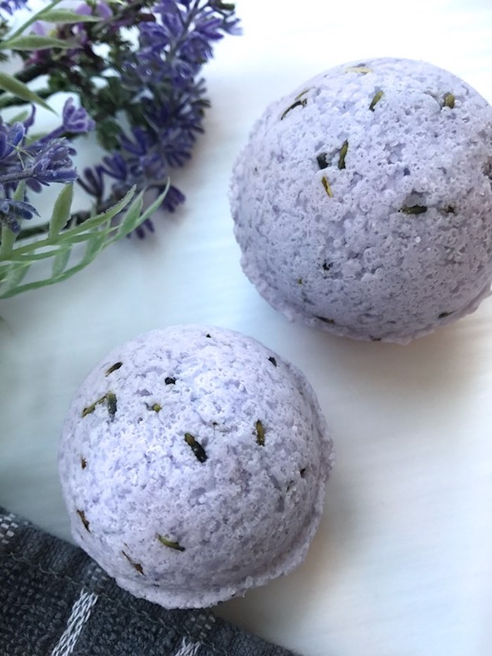 two pale violet homemade bath bombs, decorated with flower petals, on a marble surface, near a bunch of violet flowers
