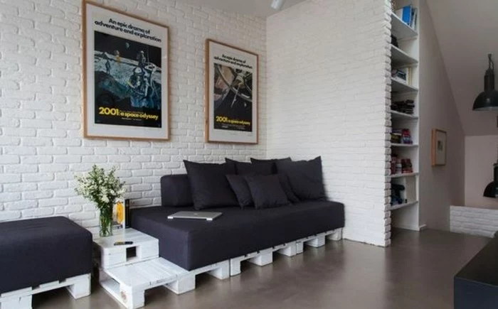brick wall in white, inside a living room with a pallet sofa, made from white wooden pallets, covered in black sofa cushions