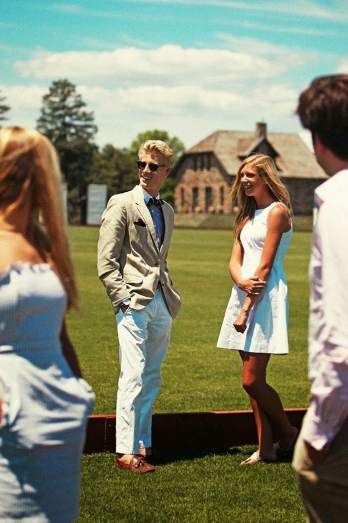 laughing couple standing on a green lawn, the man is dressed in white trousers and shirt, and beige blazer, what is cocktail attire for men, the woman sports a short white dress