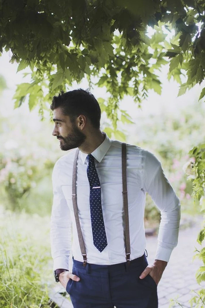 bearded brunette man, wearing a white shirt, with brown suspenders, dark patterned tie, and dark blue trousers, mens wedding guest attire, trees and grass nearby