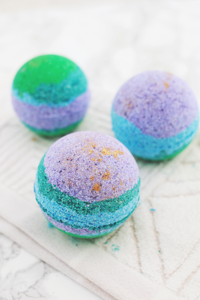 how to use a bath bomb, three spherical striped bath bombs, in green and violet, teal and orange, placed on a textured cream fabric