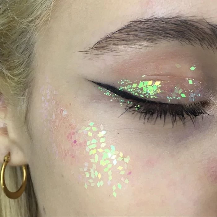 yellow and pale green, pink and silver, tiny diamond-shaped glitter pieces, decorating the eyelid and cheekbone, of a blond woman's face, make up ideas, black mascara and eyeliner