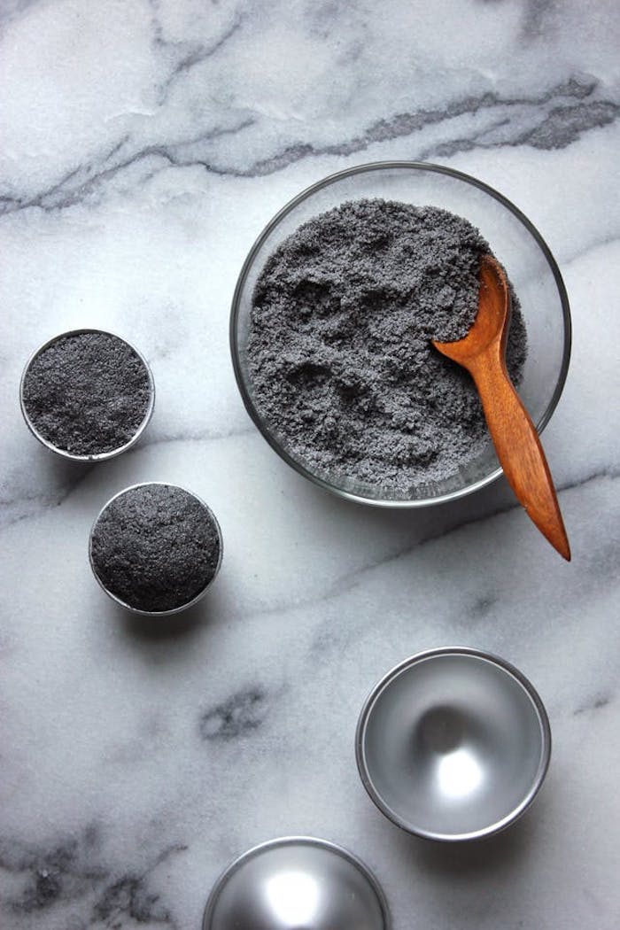 the process of making a dark grey or black bath bomb, mixing dark grey powder in a clear glass bowl, using a wooden spoon, filling a metal moulding dish with powder