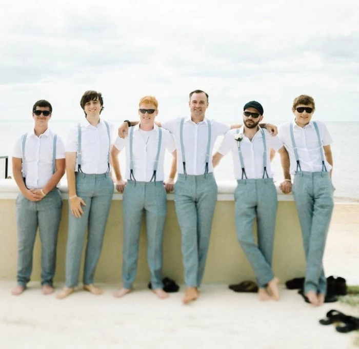 six men standing barefoot, and wearing grey trousers, white shirts and suspenders, mens casual summer wedding attire, sea in the background