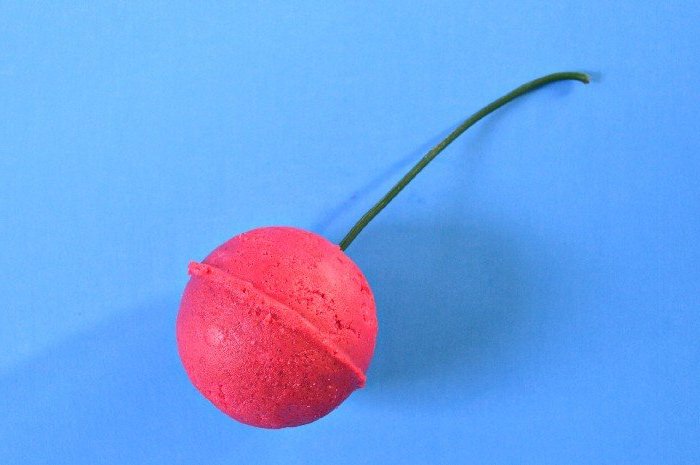 giant cherry bath bomb, dark pink in color, decorated with a long green artificial stalk, and placed on a blue surface