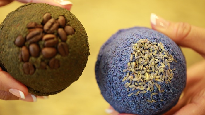 coffee beans covering a black bath bomb, held by woman's hand, with a classic french manicure, her other hand holds a textured, blue bath bomb, topped with dried lavender