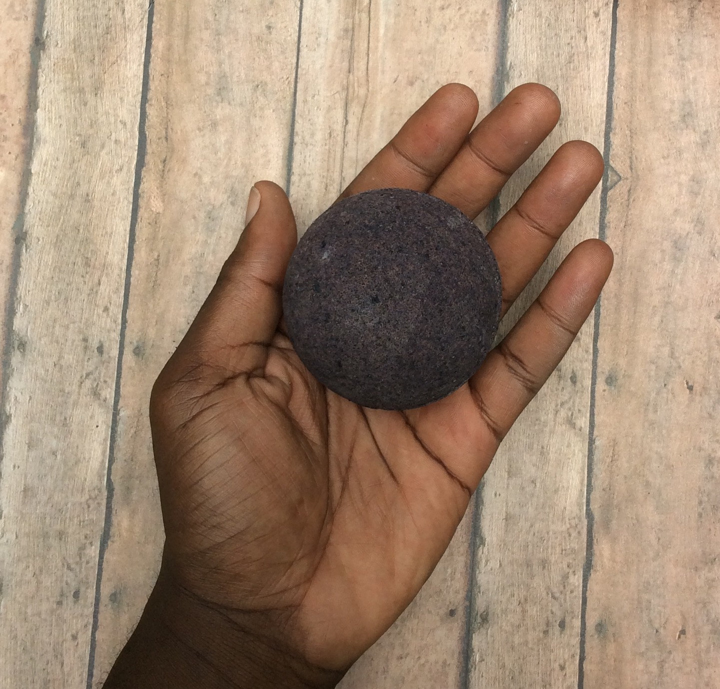 terxtured black bath bomb, in the palm of a person's hand, old worn wooden surface in the background