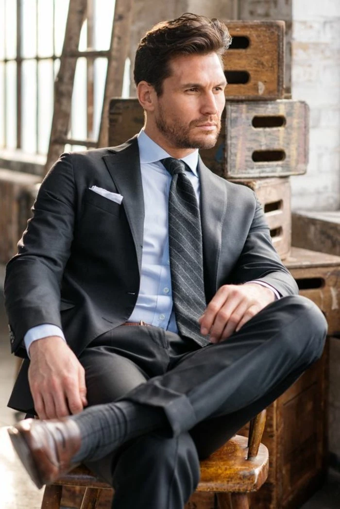 sitting brunette man, wearing pale blue shirt, dark grey two piece suit, striped dark grey tie, black tie attire for men, smart brown shoes