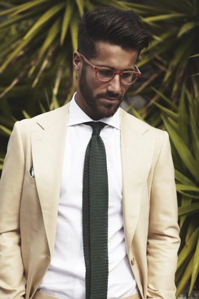 palm leaves behind a brunette man, with red-framed sunglasses, what is cocktail attire for men, ivory colored blazer, over white shirt with a dark tie