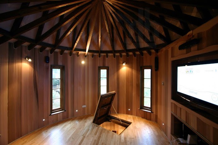 inside a tree house castle, round room with wood panelling on the walls, trap door and wooden beams on the ceiling, treehouse ideas, large TV and three windows