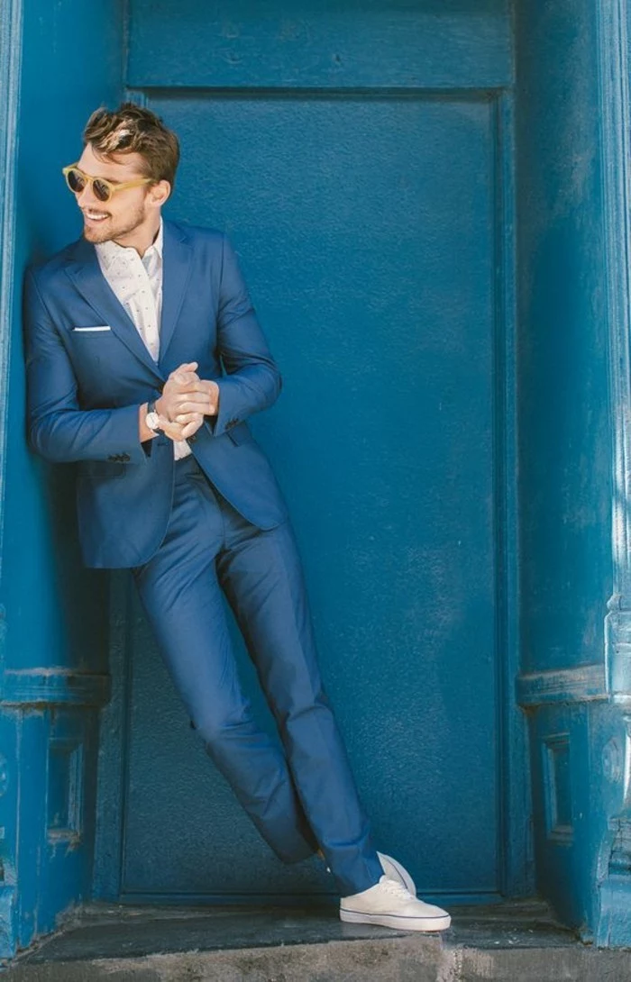 happy looking man, leaning on a blue wall, wearing a blue two piece suit, white shirt and white sneakers, mens summer wedding attire, sunglasses with yellow frames