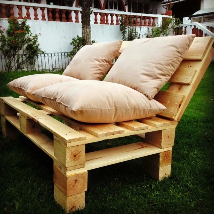garden with green grass, and a light wooden bench, made from pallets, with several beige cushions on top, pallet outdoor furniture