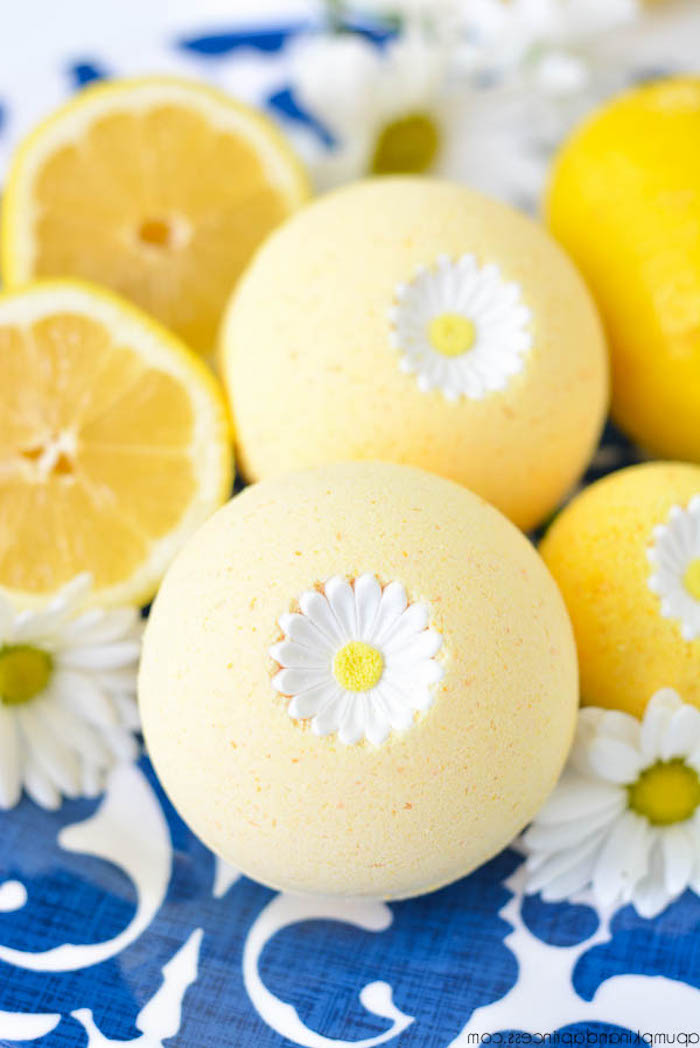 yellow bath bombs, decorated with daisies, what is a bath bomb, placed on white and blue cloth, near halved lemons and fresh daisies