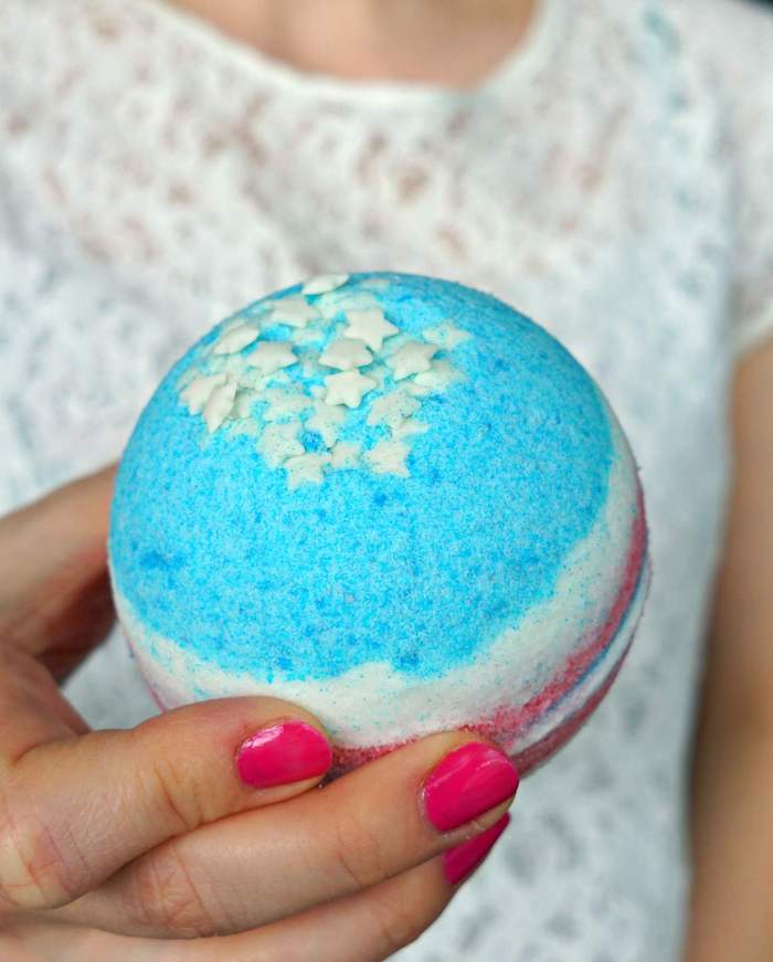 fourth of jully usa bath bomb, in pale blue red and white, with little white stars on top, festive bath balls, held by woman in white lacy top, with hot pink nail polish