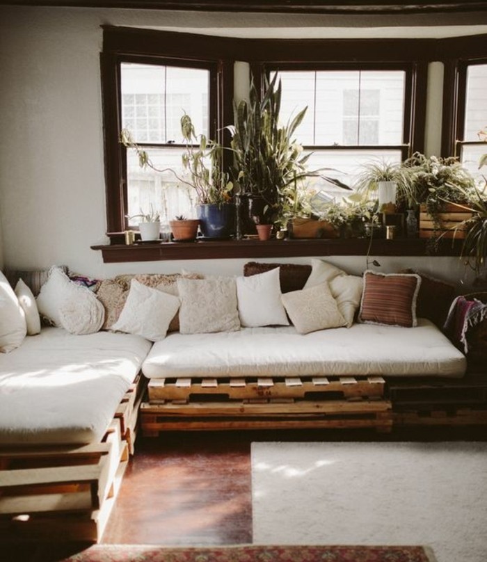 pallet sofa, covered in white, beige and brown cushions, inside a room with brown floor, pale beige rug, and a windowsill with many potted plants