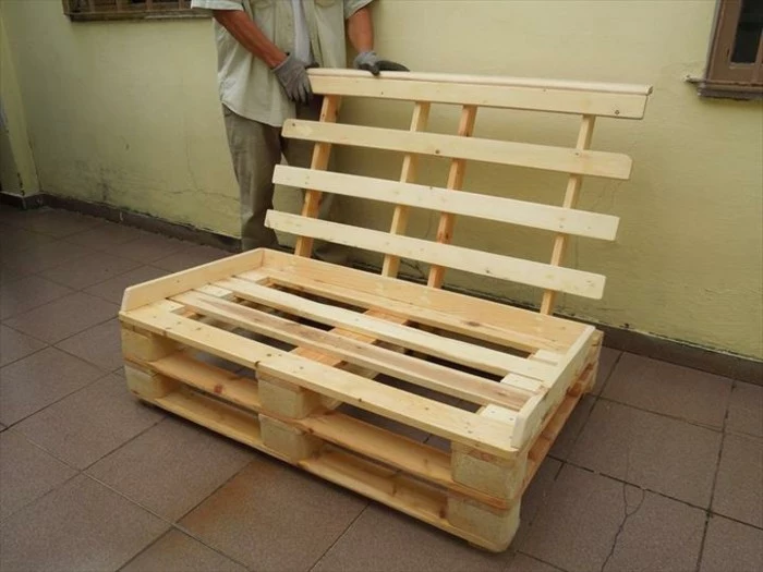 how to make pallet furniture, man in grey clothes, adding a wooden backrest, to a settee made from light wooden pallets