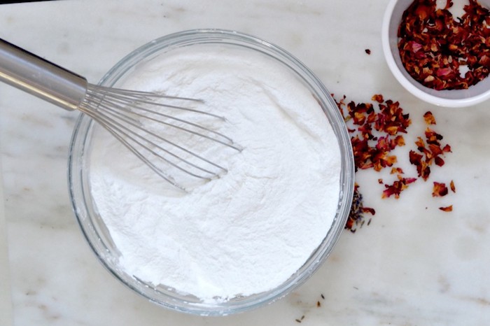 whisking fine white powder, inside a clear glass bowl, using wire mixing utensil, ingredients for bath bombs, smaller white ceramic bowl, with dried rose petals nearby