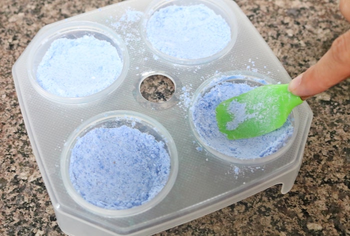 pressing blue powder, into plastic moulding dish, with four round compartments, how to make bath bombs, using green plastic spatula