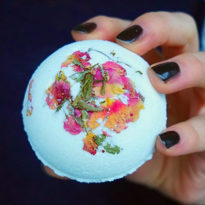 bath balls you can make at home, hand with black nail polish, holding pale blue bath bomb, decorated with dried rose leaves, and petals in yellow, orange and pink