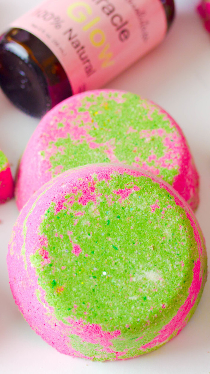 acid green and hot pink bath bombs, shaped like large round tablets, a set of two, placed on a white surface, near a bottle with pink label