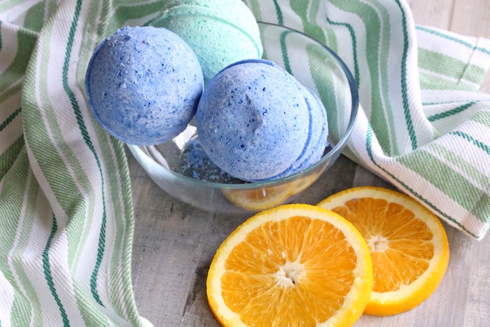 two orange slices, placed on a green and white striped table cloth, near clear glas bowl, containing two blue, and one turquoise bath bombs, how to make bath bombs