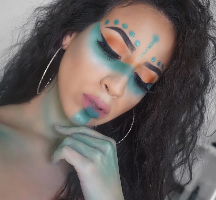 curly black hair, on young woman, wearing black eyeliner and orange eyeshadow, unicorn make up, with turquoise paint on her face, neck and hand