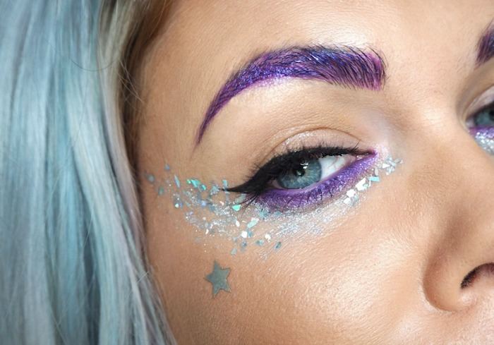 sparkly teal glitter, and purple eyeshadow, on blue eye with black eyeliner, festival makeup, woman with pale blue hair, and a purple eyebrows