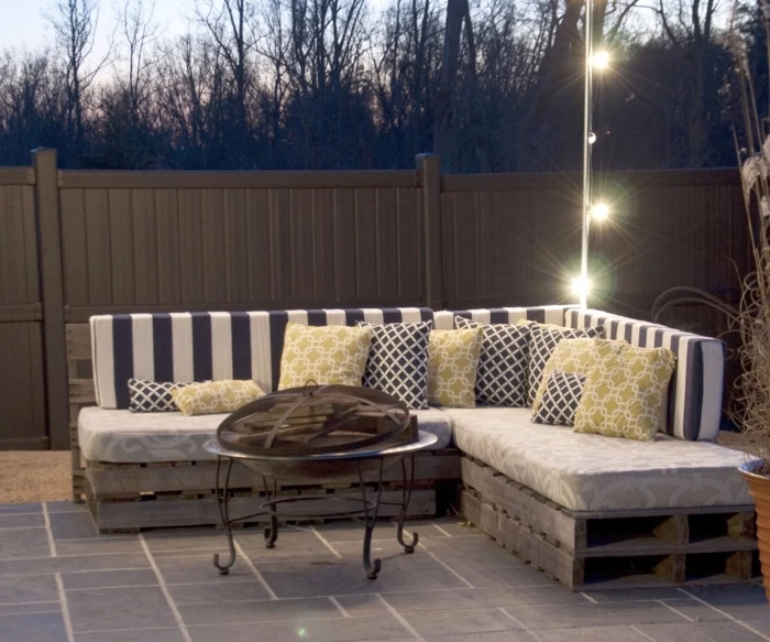 fairy lights hanging near a corner pallet settee, with soft striped backrests, and cushions with mustard, black and patterns, pallet outdoor furniture, wrought iron table