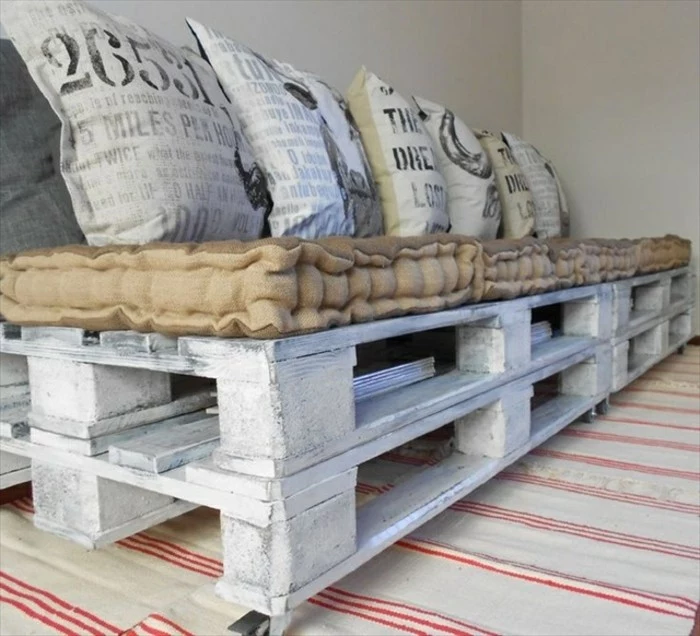 close up of a pallet sofa, made from several wooden pallets, painted in whitewash, how to make pallet furniture, beige mattress and several light grey pillows, with graphic print