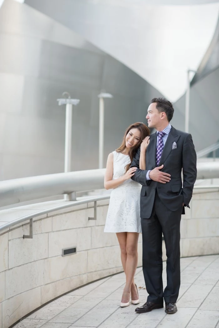 brunette east asian woman, wearing short white dress, standing hand in hand, with a man in a dark grey suit, mens summer wedding attire, wearing purple patterned tie