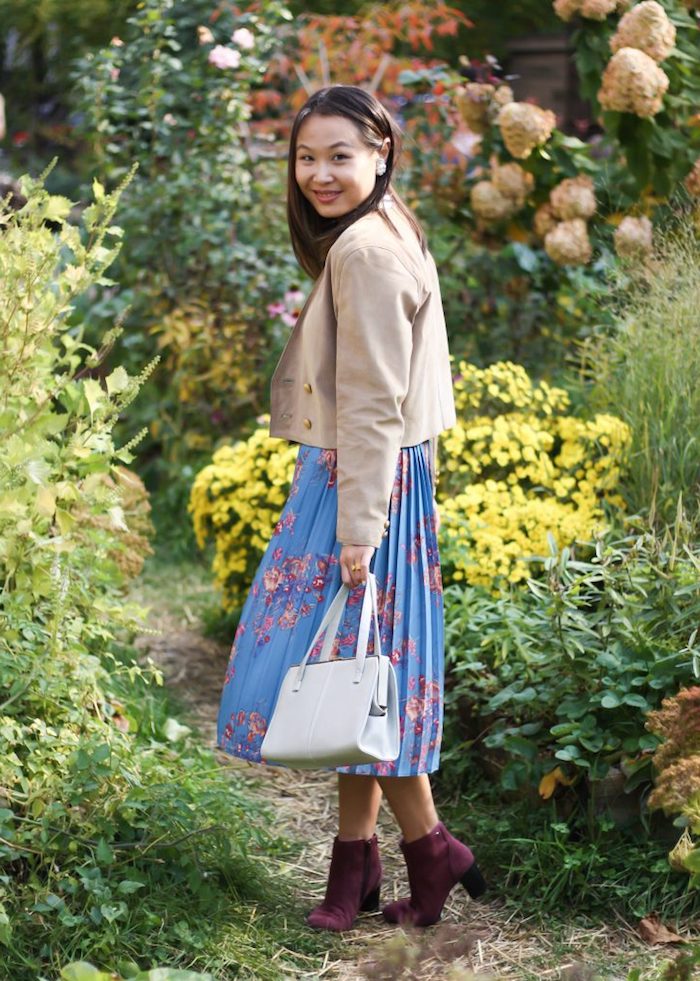 smiling brunette girl, wearing a blue pleated skirt, with pink floral pattern, purple ankle boots, and a beige jacket, garden party attire, white leather hand bag