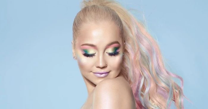 smiling young woman, with platinum blonde hair, featuring strands of pastel blue, pink and purple, tied in a ponytail, wearing unicorn makeup, with green and yellow eyeshadow
