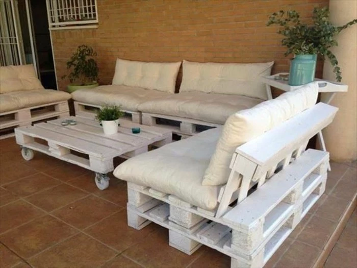 a set of three white sofas, made from pallets covered in pillows, and a matching white pallet table on wheels, how to make pallet furniture, placed on a porch