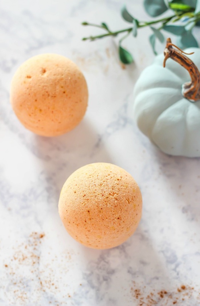 orange bath bombs, placed on a marble surface, how to use a bath bomb, near a small, pale green pumpkin, and a branch with green leaves