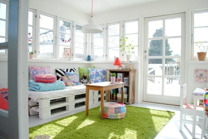 diy sofa made from white pallets, and decorated with multicolored cushions, inside a bright room, with several windows and a glass door