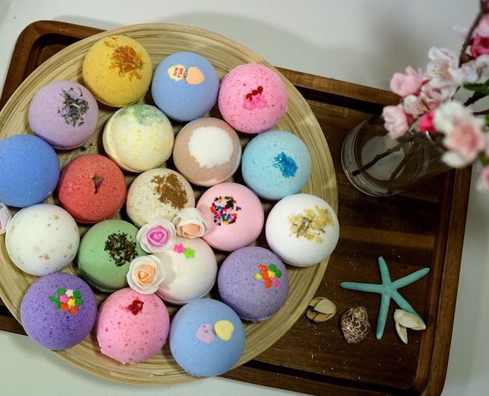 assortment of bath bombs, in different colors, and with various decorations, on a round tray, placed on a rectangular tray, near flowers and shells