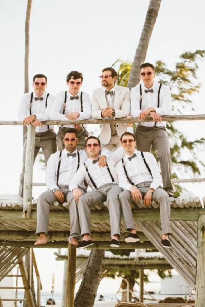 groom in white suit, surrounded by six barefoot men, wearing matching clothes, mens wedding guest attire, white shirts and beige trousers, with suspenders and bowties