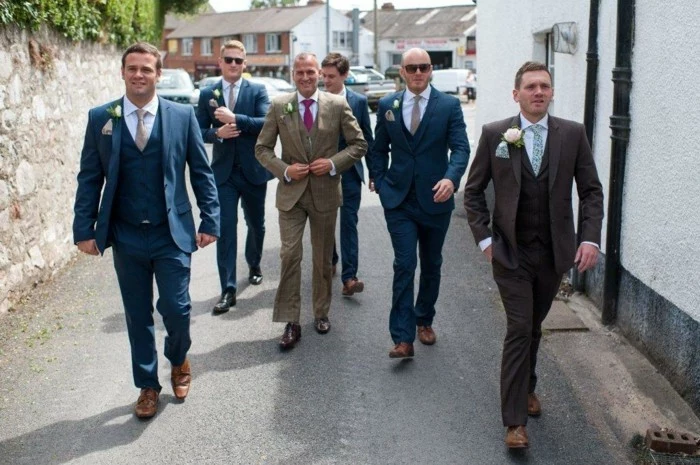 group of men in dark blue, and beige suits, with ties in different colors and boutonnieres, how to dress for a wedding male, walking down the street