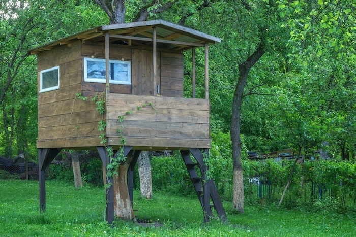 classical small backyard treehouse, made of wood, on metal platforms, with two small windows, and a metal ladder, green trees nearby