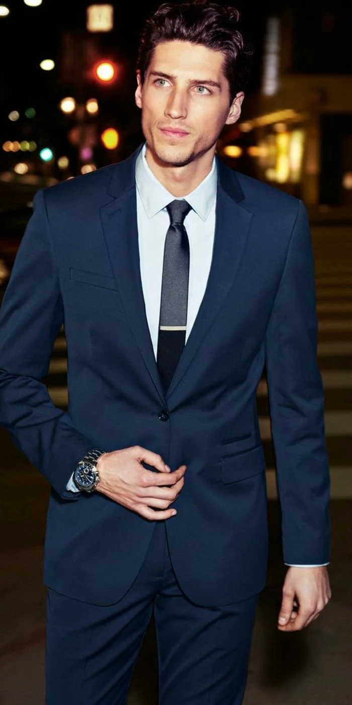 young man wearing a dark blue, two piece suit, with white shirt, and a shiny black tie, with a silver pin, black tie attire for men, a street at night