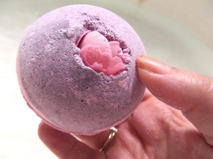 hand with a ring, holding a a pale purple bath bomb, decorated with pink soap fragments, what is a bath bomb, spherical shape