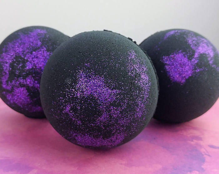 three black bath bombs, decorated with sparkly dark purple glitter, and placed on a pinky-purple surface