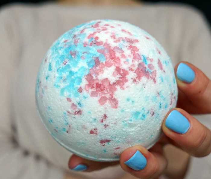 round white bath ball, decorated with pale blue and pink specks, held by hand, with pale blue nail polish, and seen in close up
